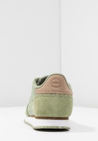 Baskets en suède vert et tissu avec semelle blanche, patch en cuir marron embossé du mot "WODEN" au talon, vues de dos sur une surface blanche.