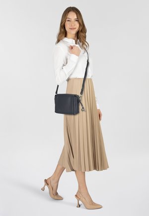 Borsa a tracolla in pelle nera con spallaccio regolabile, caratterizzata da una finitura liscia e una chiusura con zip. Il modello indossa una camicia bianca e una gonna plissettata beige.