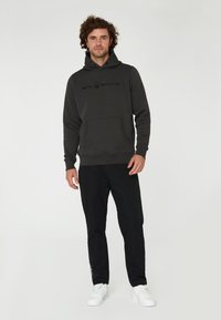 Sudadera gris oscuro con un bolsillo frontal, presenta un logotipo discreto en el pecho, combinada con pantalones negros y zapatillas blancas, con una textura suave.