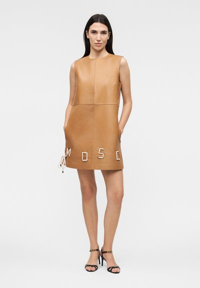 Γυναίκα που φοράει αμάνικο δερμάτινο φόρεμα σε ταμπά χρώμα με μπροστινές τσέπες και μαύρα σανδάλια με ψηλά τακούνια, στέκεται μπροστά σε απλό φόντο.