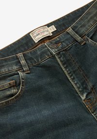 Tætsiddende mørkeblå denimjeans med kobberstin, en frontknap og et mærke med teksten "NEXT Denim Goods Co" nær taljen.