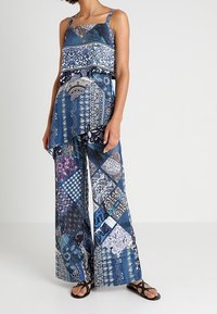 Femme portant un haut sans manches à motif patchwork bleu et un pantalon large assorti avec des sandales noires sur fond uni.
