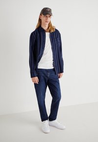 Granatowa koszula w paski z guzikami, biała koszulka pod spodem, dżinsy oraz białe sneakersy. Szary czapka dopełnia stylizację.