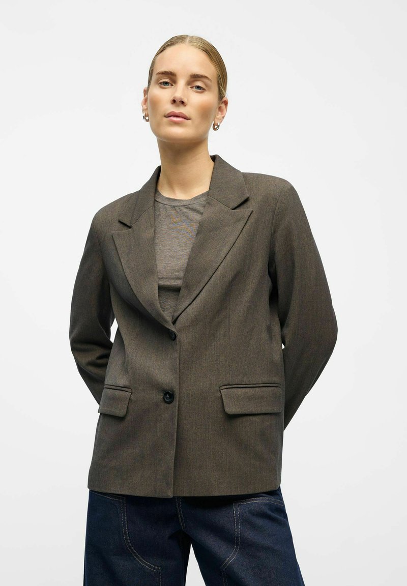 Blazer marrone su misura con rever affilati, chiusura a un bottone e due tasche frontali, indossato sopra un top grigio su uno sfondo neutro.