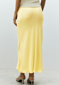 Jupe maxi en satin jaune avec une texture lisse, présentant une légère évasement et une fente sur le côté. Portée avec des sandales à talons beiges.