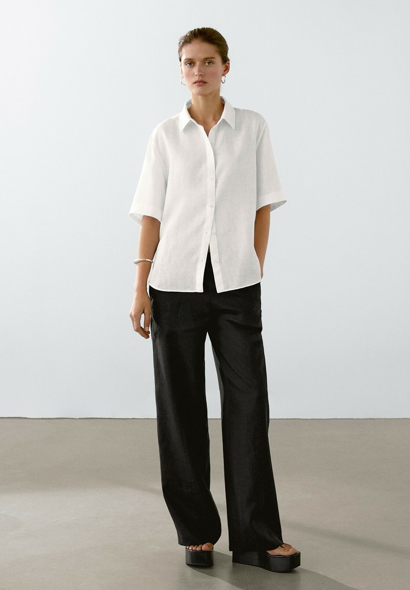 Camicia bianca a maniche corte, abbinata a pantaloni neri a gamba larga e sandali platform spessi. Tessuto lucido con design minimalista.