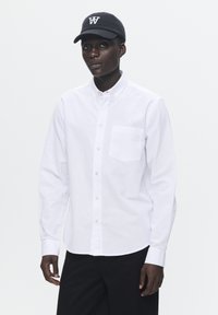 Camisa blanca de botones con mangas largas, bolsillo frontal y bordado de un pequeño logo. Combinada con una gorra negra que tiene un logo blanco.