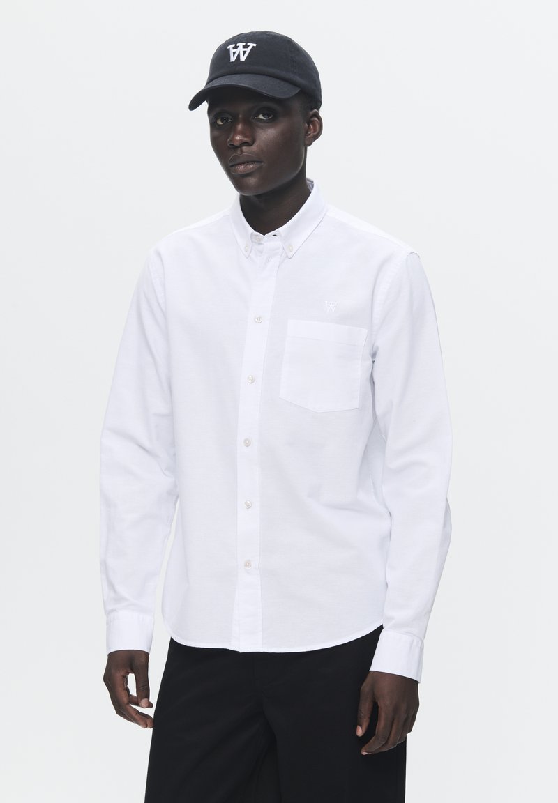 Camisa blanca de botones con mangas largas, bolsillo frontal y bordado de un pequeño logo. Combinada con una gorra negra que tiene un logo blanco.