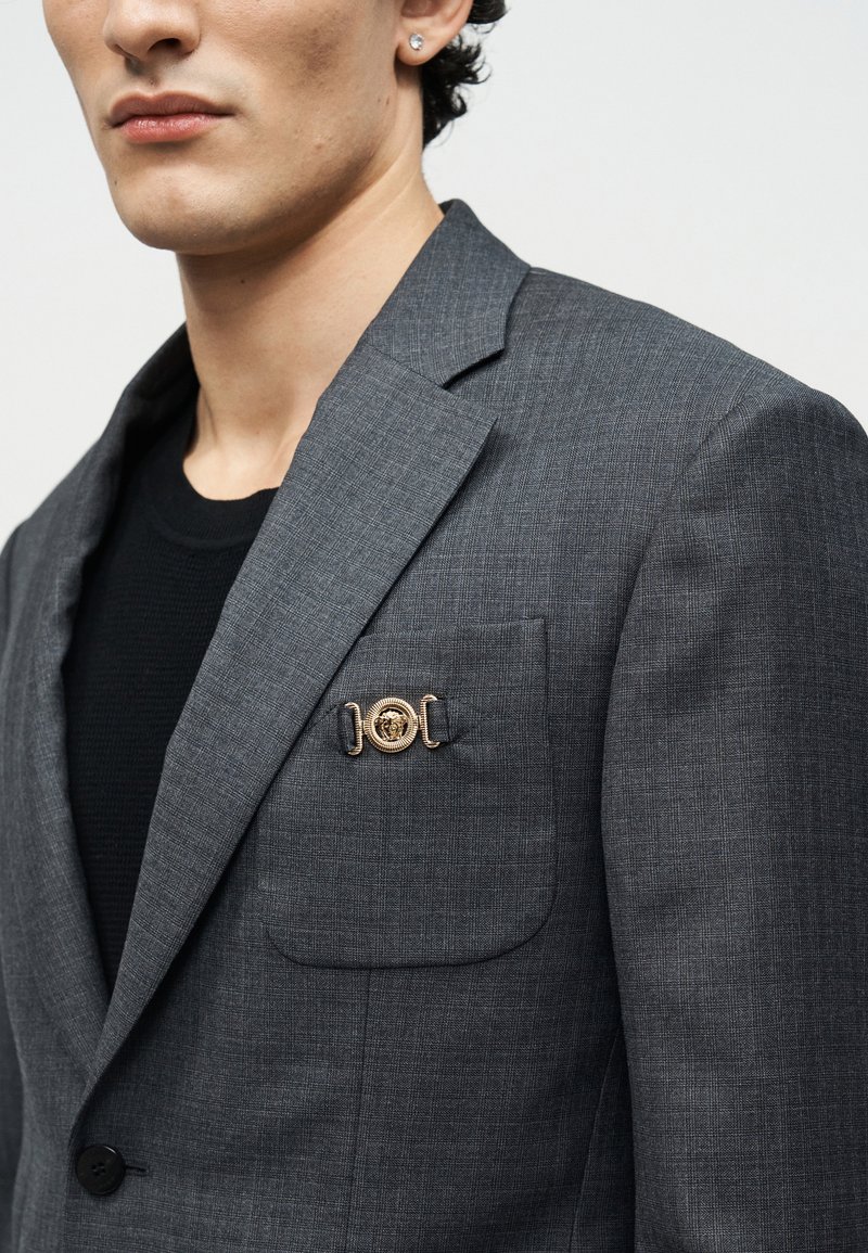 Homme portant un blazer gris foncé à carreaux avec une broche décorative dorée sur la poche poitrine et une petite boucle d'oreille clou.