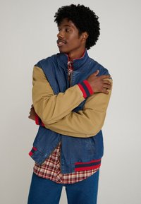 Denim bomber jacket with tan sleeves, red-striped cuffs, and zippered closure. Layered over a red plaid shirt with blue jeans.
