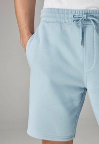 Pantaloni corti in cotone azzurro chiaro con vita elastica e cordoncino, dotati di tasche laterali e una texture liscia. L'orlo si trova sopra il ginocchio.