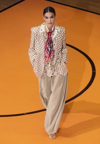 Beige rutigt kavaj med guldknappar, lager på lager över en prickig skjorta. Matchas med löst sittande beige byxor och en färgglad silkesscarf.