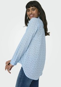 Camicia a maniche lunghe blu chiaro con piccoli pois a contrasto, colletto con bottoni e orlo curvo. Indossata con jeans scuri.