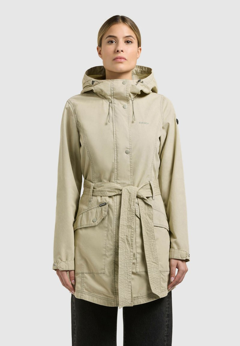 Femme portant un manteau beige à capuche avec une ceinture à la taille, des poches avant et des boutons, se tenant devant un fond gris clair uni.