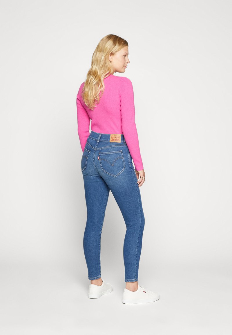 Jeans skinny blu con vita alta, caratterizzati da un'etichetta in pelle sul retro. Il modello indossa un maglione aderente di colore rosa brillante e scarpe bianche.