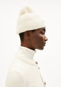 Witte gebreide muts met een ronde top, gemaakt van zachte garen. Model draagt een witte coltrui en een crèmekleurige cardigan met bruine knopen.
