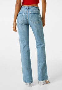 Vaqueros de mezclilla azul claro de campana con cintura alta, cinco bolsillos y un acabado texturizado. Combinados con un top corto rojo y tacones transparentes.
