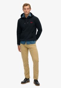 Felpa della Marina con tasca frontale, cappuccio con cordino e logo rosso. Camicia di jeans azzurra chiara sotto, abbinata a pantaloni khaki e scarpe nere.