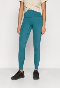 Leggings taille haute couleur sarcelle avec une texture lisse et des détails de coutures discrètes, assortis d'un haut beige clair à nouer sur le devant et de baskets noires.