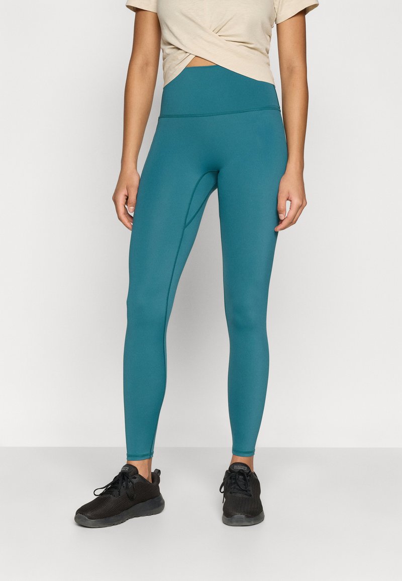 Leggings taille haute couleur sarcelle avec une texture lisse et des détails de coutures discrètes, assortis d'un haut beige clair à nouer sur le devant et de baskets noires.