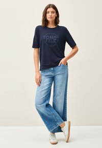 T-shirt en coton bleu marine à manches courtes et col rond avec impression de texte blanc. Associé à un jean en denim bleu clair à jambes larges et des baskets blanches.