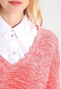Maglioncino rosa corallo a maglia con scollo a V, caratterizzato da una texture a costine e un tessuto morbido, indossato sopra una camicia bianca con colletto e bottoni argentati.