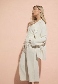 Ljusbeige linnekostym i tre delar bestående av en avslappnad blazer, en knäppt väst och vida byxor med framfickor och minimal detaljering.