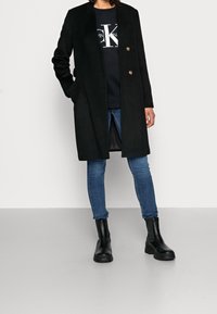 Abrigo de mezcla de lana negro con corte recto, dos acentos de botones dorados y bolsillos laterales, que se lleva sobre una sudadera negra y unos vaqueros azules.