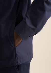 Camicia blu navy con una texture di tessuto liscia, dotata di polsini con bottoni e tasche laterali, parzialmente visibile su uno sfondo neutro.