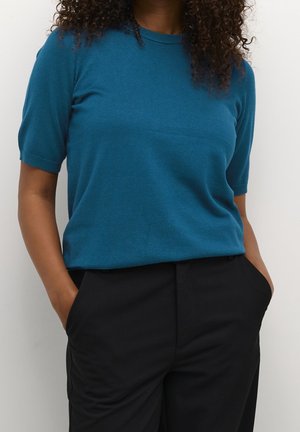 Femme aux cheveux bouclés portant un haut à manches courtes bleu canard et un pantalon noir, debout les mains dans les poches devant un fond uni.