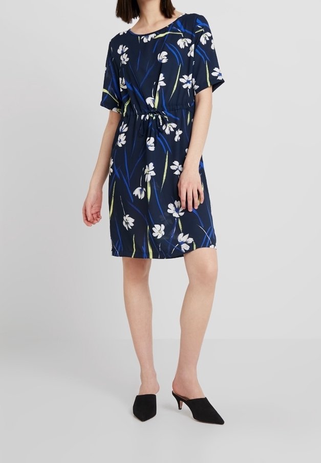 Femme portant une robe bleu marine longueur genou avec un imprimé floral blanc, des manches courtes, et des chaussures noires à talons sans lacets, debout sur un fond uni.