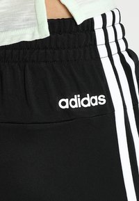 Pantalon de survêtement Adidas noir avec taille élastique et trois bandes blanches sur le côté, porté avec une chemise de couleur claire.