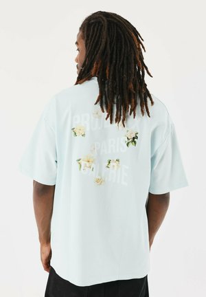 Camisa de manga corta azul claro con estampado floral y texto blanco en la parte de atrás. Fabricada con tela suave, presenta un ajuste relajado y un escote redondo.