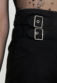 Černé kalhoty s vysokým pasem vyrobené z texturované látky, s dvojitými stříbrnými sponami a hladkým páskem. Průhledný síťovaný top s dekorativními flitry.