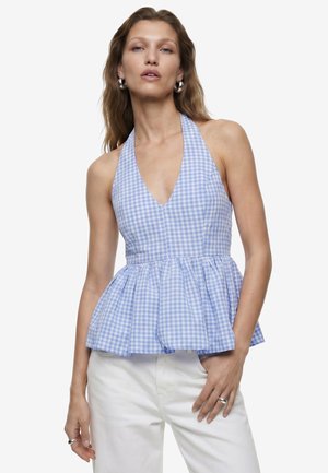 Femme portant un top halter sans manches à carreaux vichy bleu et blanc avec un bas à volant peplum et un pantalon blanc, debout avec une main dans la poche.