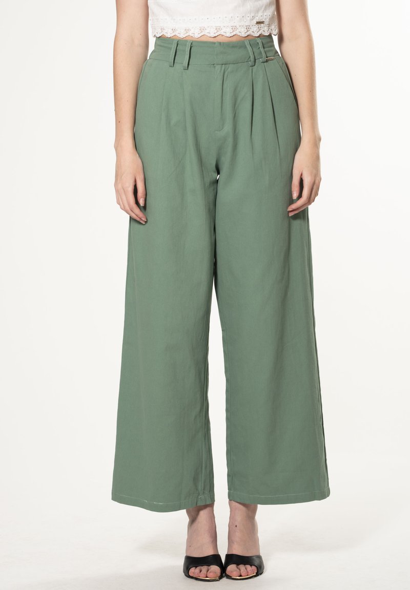 Brede broek in zachte groene stof, met plooien, een hoge taille en riemlussen. Gecombineerd met zwarte sandalen met hakken.