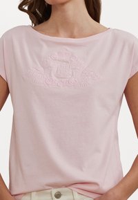 Camiseta de algodón rosa claro con escote ancho, mangas cortas y detalle bordado de un crest floral en el centro. Textura suave y ajuste relajado.