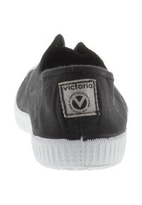 Zapatilla de lona negra con suela de goma blanca; presenta un parche con logo en la parte trasera, forma redondeada y suela texturizada. Adecuada para uso informal.