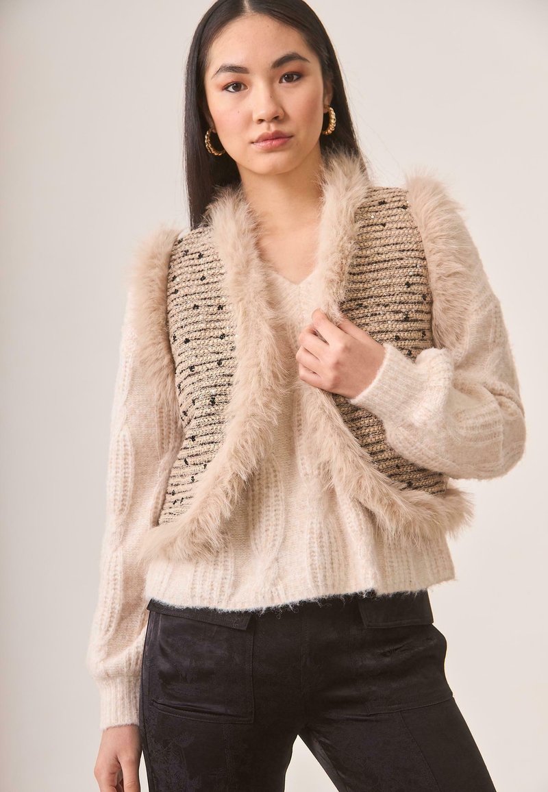 Suéter de punto grueso en crema con detalles de piel sintética beige y chaleco tejido con manchas negras, combinado con pantalones ajustados oscuros.