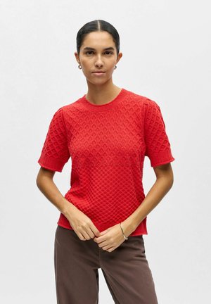 Femme aux cheveux foncés portant un pull rouge texturé à manches courtes et un pantalon marron, debout devant un fond clair uni.