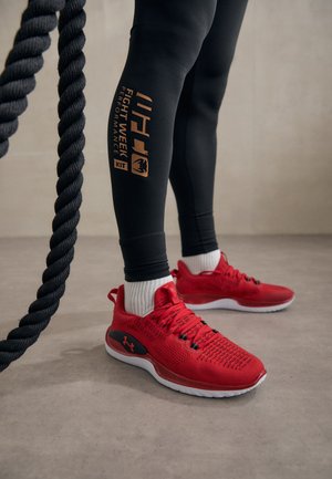 Czerwone buty sportowe z czarnymi akcentami, teksturowana siateczka u góry i biała podeszwa. Czarne legginsy z złotym detalem graficznym i grubym sznurem w tle.