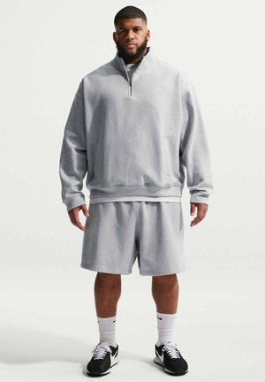 Mann trägt hellgrauen Sweatshirt mit Viertel-Reißverschluss und Shorts, weiße Socken und schwarz-weiße Sneakers, steht vor schlichtem weißen Hintergrund.