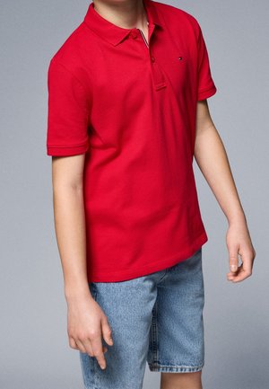 Dítě oblečené v jasně červené polokošili se dvěma knoflíky, doplněné světle modrými džínovými šortkami na šedém pozadí.