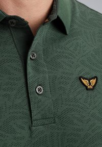 Zelena polo majica s teksturiranim uzorkom, dva crna gumba i žutim izvezenim logotipom na prsima. Kratak ovratnik i mekana tkanina.