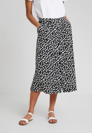 Femme portant une jupe midi noire avec un imprimé floral blanc et des boutons sur le devant, associée à des sandales blanches et un haut blanc à manches courtes.