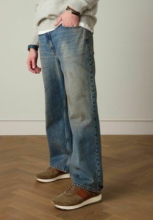 Man die losse blauwe spijkerbroek, beige sneakers, een grijze trui met armbanden en een smartwatch draagt, staand op een houten parketvloer binnen.