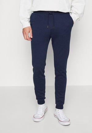 Pantalones de chándal de algodón azul marino con cintura ajustable, puños ajustados y dos bolsillos laterales, combinados con zapatillas blancas. Textura suave.