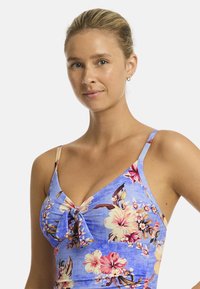 Model met blond haar in een bloemrijke blauwe outfit, poseert zelfverzekerd met een zachte glimlach tegen een effen witte achtergrond.