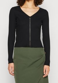 Top lungo a maniche ribattute nere con scollatura a V e zip frontale. Design aderente con una lunghezza cropped, abbinato a una gonna verde.