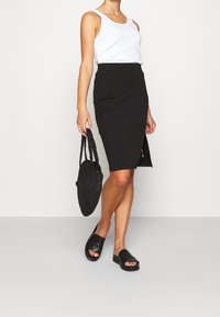 Jupe crayon noire avec une fente sur le côté, associée à un débardeur blanc et des sandales plates noires. Les accessoires comprennent un sac rond tressé noir.